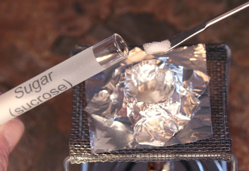 sucrose on spatula coming from test tube being put on aluminum crucible